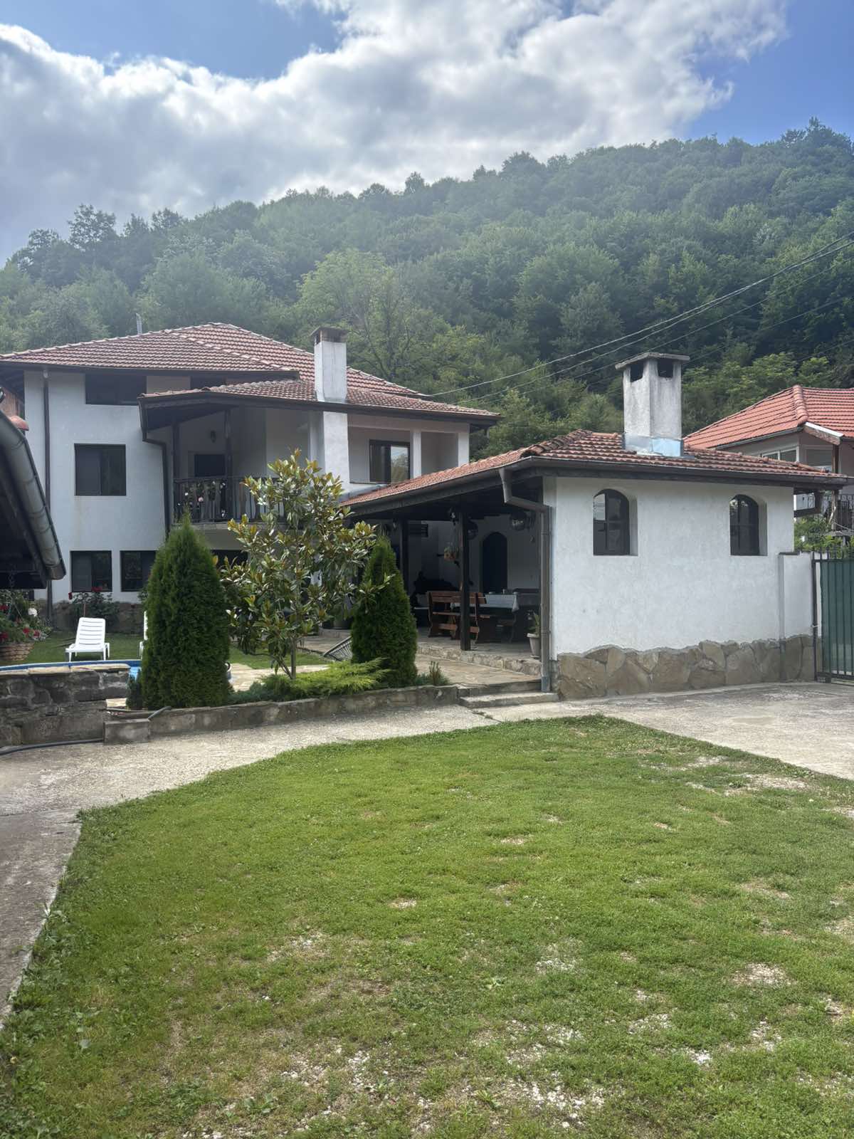 Фасадата на Йовковата Къща откъм двора: бяла къща с дървен балкон и зелена морава